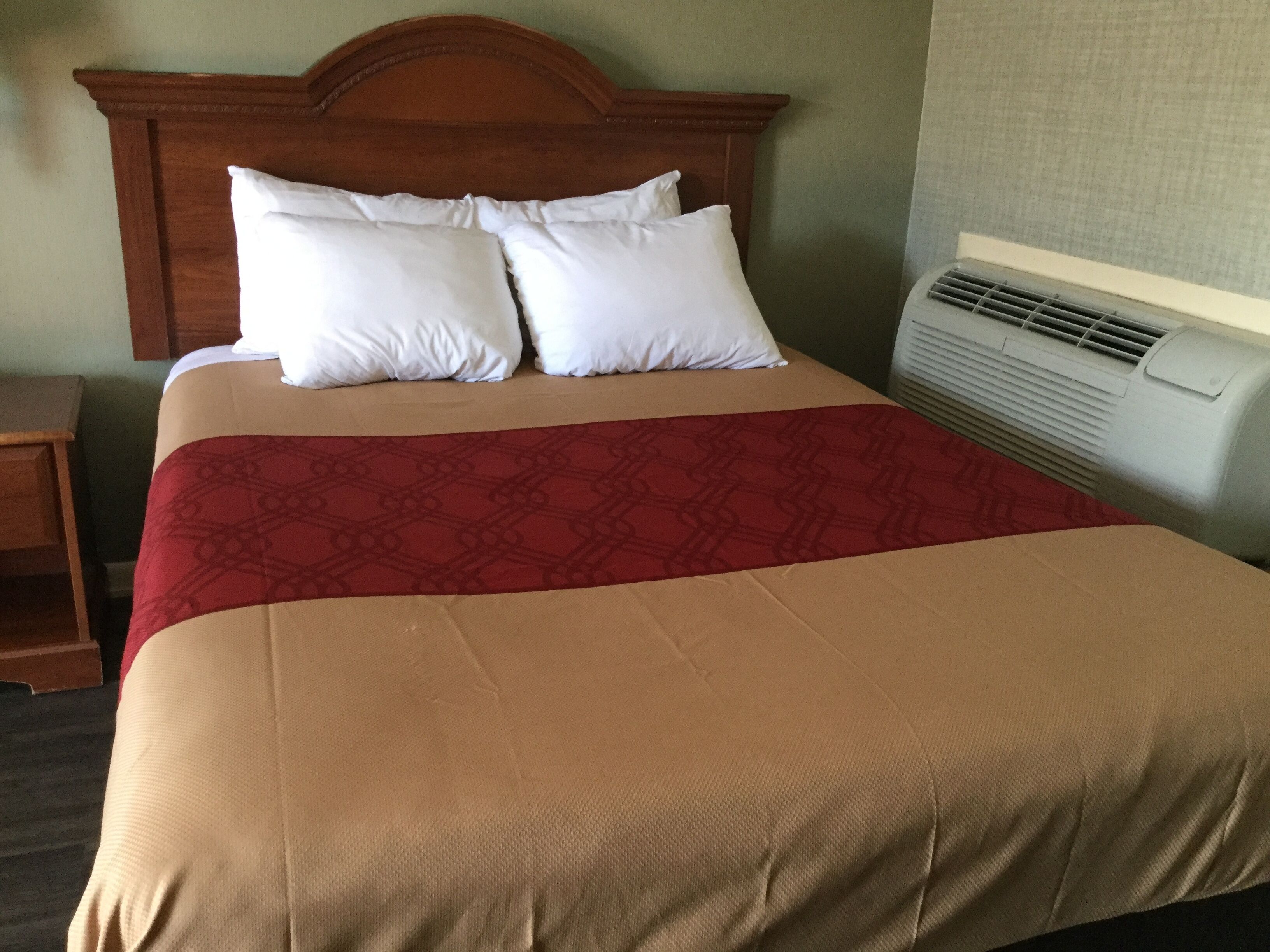 Quarto de hotel com uma cama grande, cabeceira de madeira, duas almofadas brancas, edredom vermelho e bege, e ar condicionado fixado na parede.