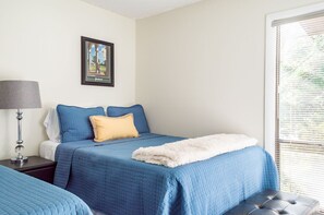 A bedroom with a bed, a nightstand, a lamp, a window with blinds, and a framed picture on the wall.