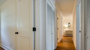 Un pasillo con puertas y contraventanas blancas, suelo de madera, y una pequeña mesa con una lámpara y objetos decorativos al fondo.