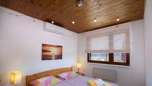 A bedroom with a wooden ceiling, a bed with colorful pillows, a nightstand with a lamp, and a window with blinds. - House Petra on the beach-nice view (Cesarica)