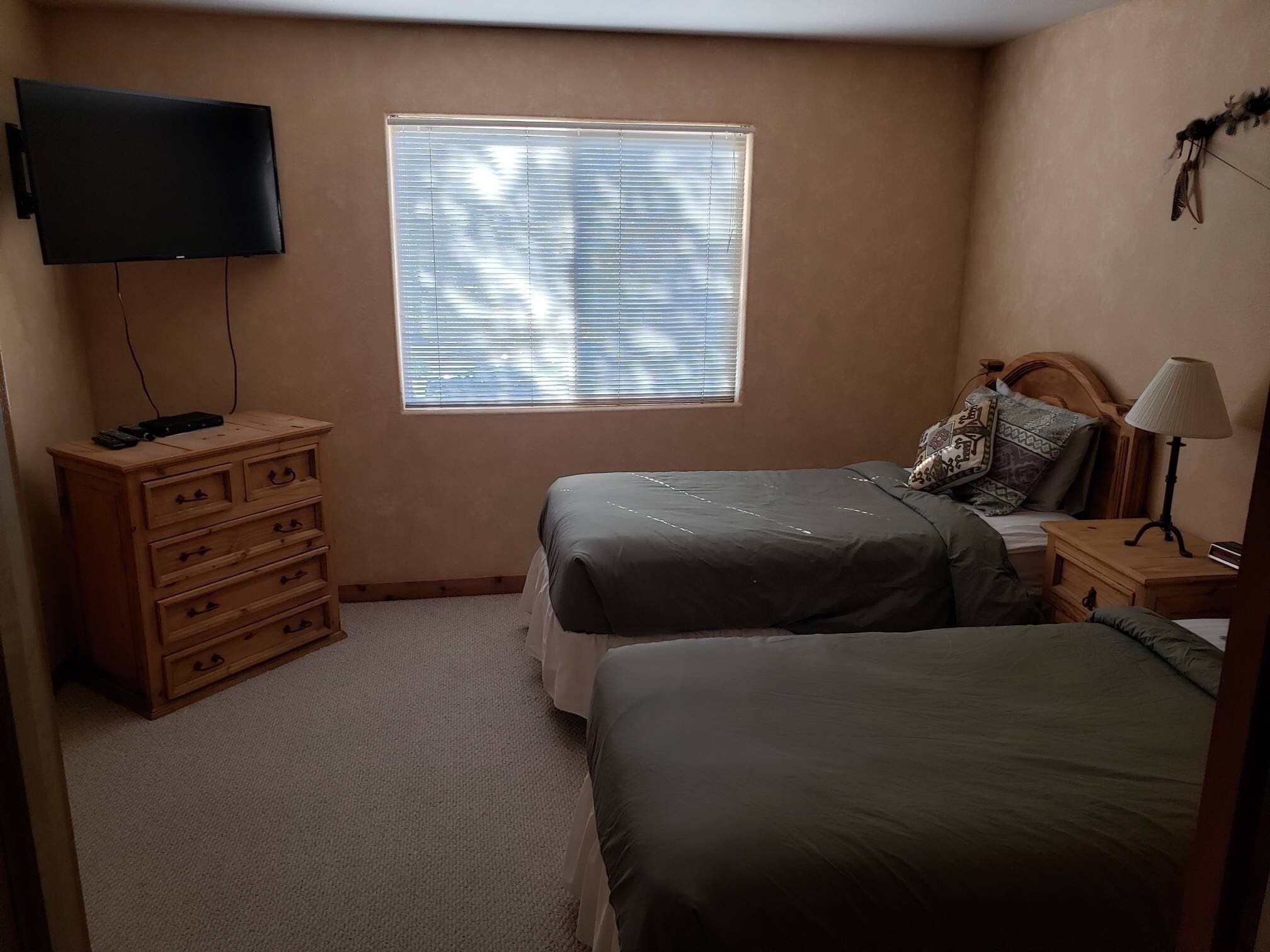 Ein Schlafzimmer mit zwei Betten, einem Schminktisch, einem Fernseher, einem Fenster mit Jalousien, einer Lampe und einem Nachttisch.