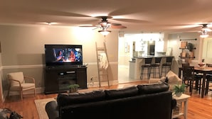 Een woonkamer met een zwarte leren bank, een televisie en een plafondventilator. Achtergrond is een keuken.