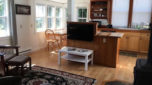 A spacious living room with a TV, wooden furniture, a dining area, and a kitchen with wooden cabinets and a window.