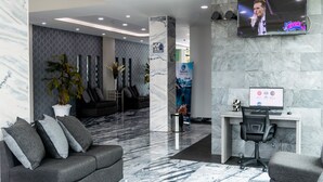 A modern lobby with a marble floor, a sofa with cushions, a desk with a computer, and a TV displaying a person. - Hotel 5 Inn Plus (León)