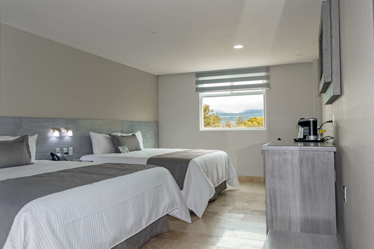 A hotel room with two beds, a small desk, a coffee machine, and a window with a view of the mountains.