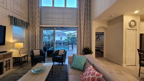 A spacious living room with a large sofa, two armchairs, a coffee table, and a television. There is a balcony with a view of the outside.