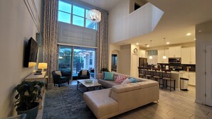 A spacious living area with a sofa, coffee table, and a dining area with chairs. There is a kitchen with modern appliances and a large window providing natural light.