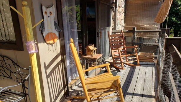 Una terraza de madera con sillas mecedoras y una pequeña mesa, una decoración de búho de madera y un frasco colgado en la barandilla.