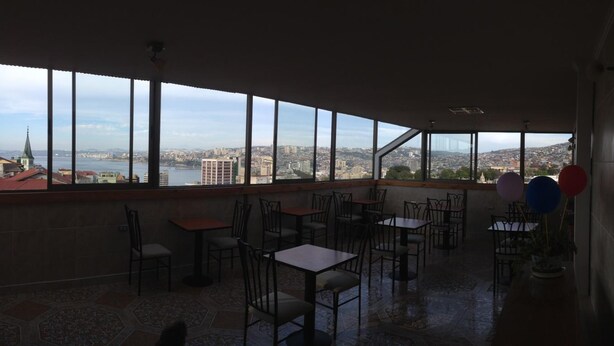 Un restaurante con amplios ventanales que ofrecen vistas a la ciudad, mesas y sillas dispuestas para comer, y algunos globos a la derecha.