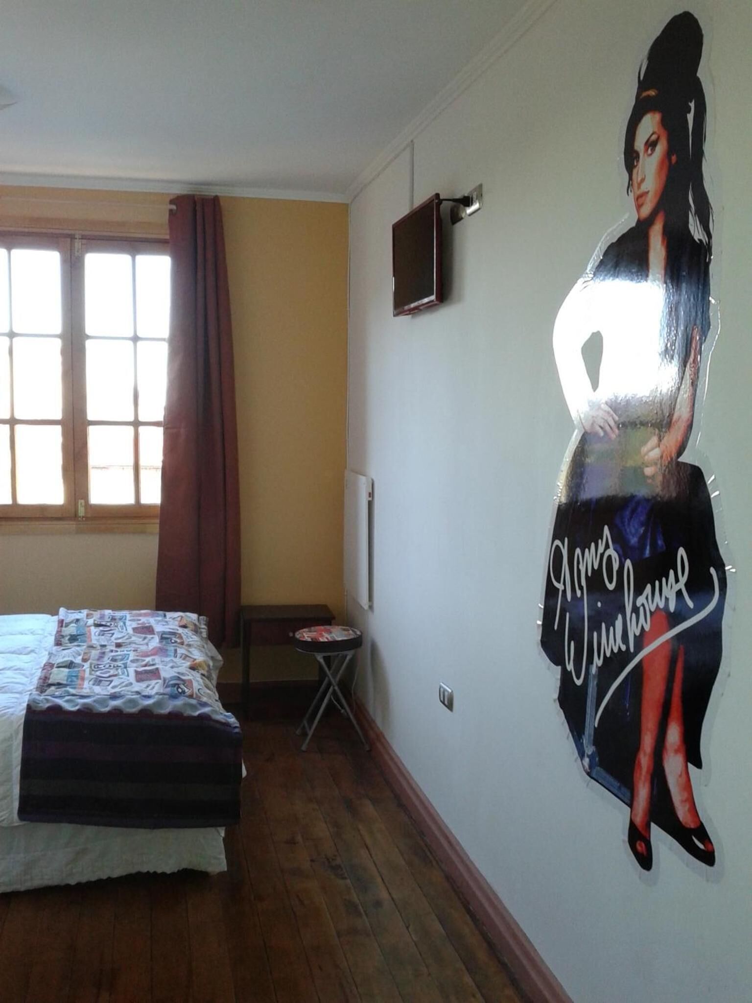 A bedroom with a bed, a small table, a TV on the wall, and a large poster on the wall.
