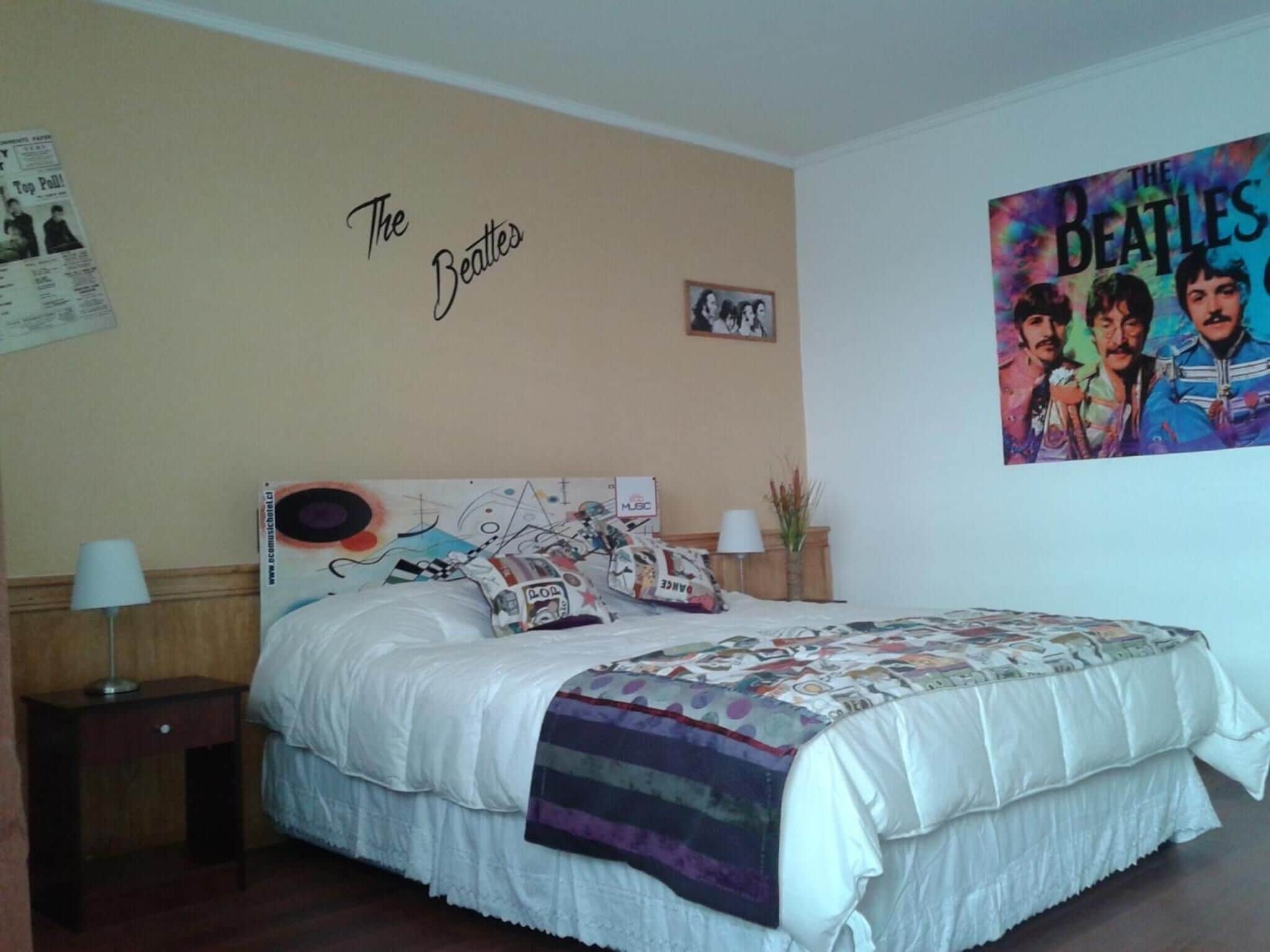 Et soveværelse med en seng, et vægur, et rammet billede og et plakat af The Beatles.