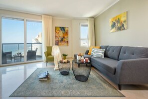 A modern living room with a grey sofa, two black wireframe coffee tables, a green armchair, and a large window with a view of a cityscape and a cruise ship.