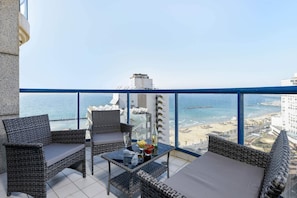Ein Balkon mit Korbmöbeln, einem Glastisch und Blick auf Strand und Meer.