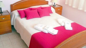 A neatly made bed with pink pillows and a white blanket, a bedside table with a lamp, and a window with sheer curtains.