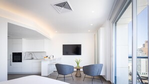 Quarto de hotel moderno com uma cama grande, área de jantar com duas cadeiras, cozinha com armários brancos, televisão de tela plana e vista para a cidade.