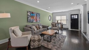 Un salon comprenant un canapé, un fauteuil, un pouf et une table basse. On y trouve également une table à manger avec des chaises, une fenêtre avec des stores et une porte.