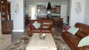 A spacious living room with a brown leather sofa, a coffee table, and a rug. There is a kitchen in the background with wooden cabinets and a black range hood. - Private Home & Fenced POOL 32'x16', sleeps 15, Jacuzzi,  TV/Game rm, Play-Set (La Verkin)