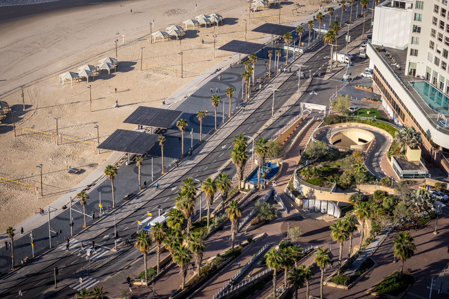 Vue aérienne d’une zone côtière avec une route, des palmiers et un bâtiment à la façade incurvée.