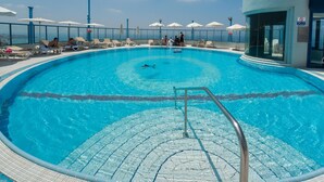Una piscina circular con agua azul cristalina, rodeada de sillones de descanso y sombrillas, y un edificio con paredes de vidrio a la derecha.