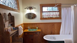Een badkamer met een houten wastafelmeubel, een vrijstaand bad, een raam met gordijn en een spiegel boven de wasbak.