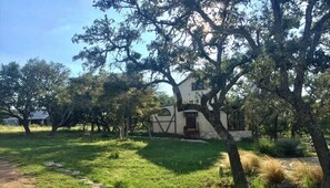 Un bâtiment de style rustique, doté d’une façade blanche et de moulures sombres, entouré d’une végétation luxuriante et d’arbres.