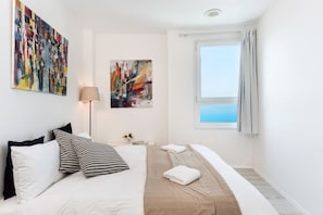 Habitación de hotel con cama, dos cuadros, una ventana con cortinas y vista al mar.