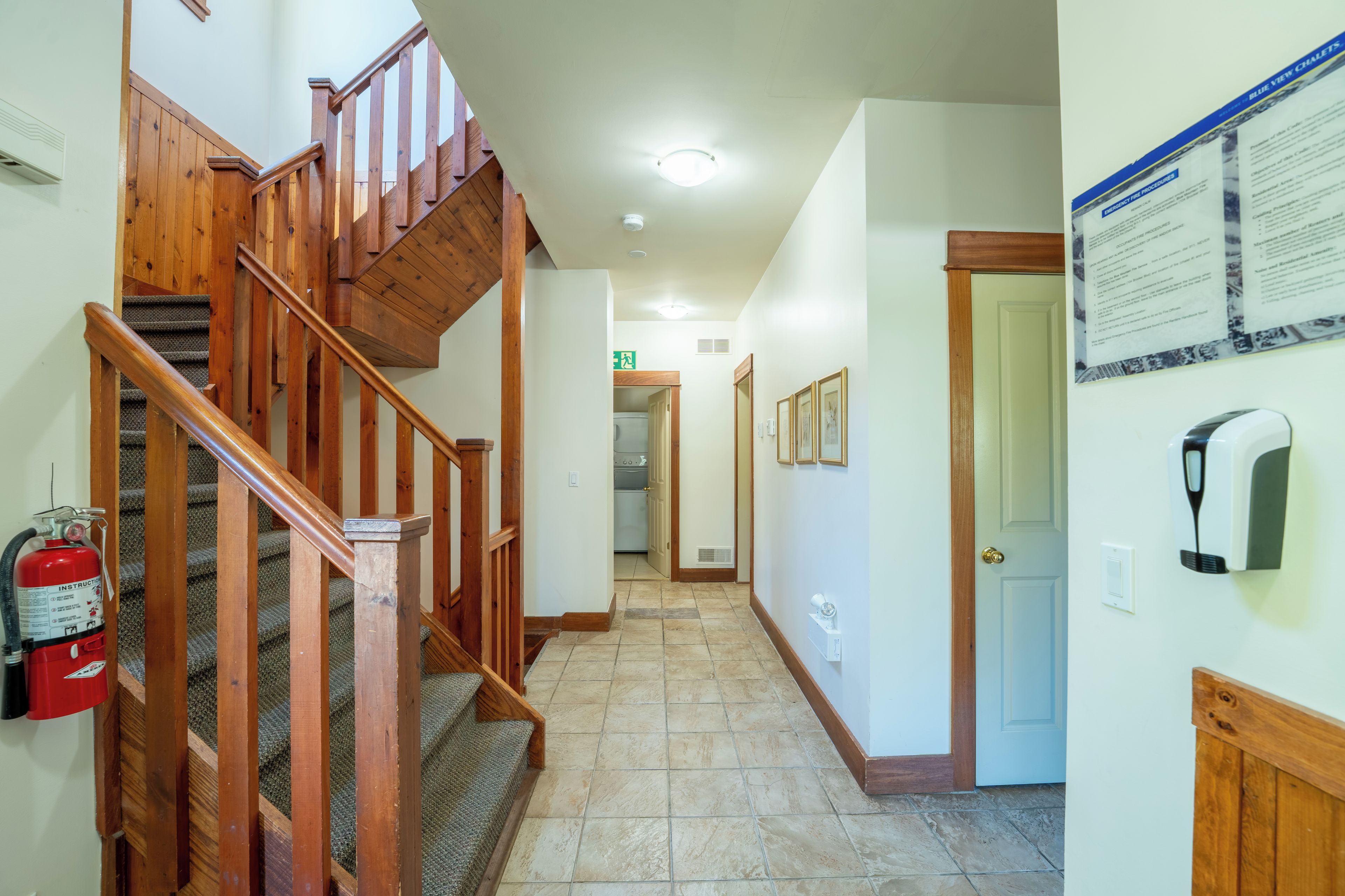 A hallway with a wooden staircase, a fire extinguisher, a hand sanitizer dispenser, and a door with a sign.