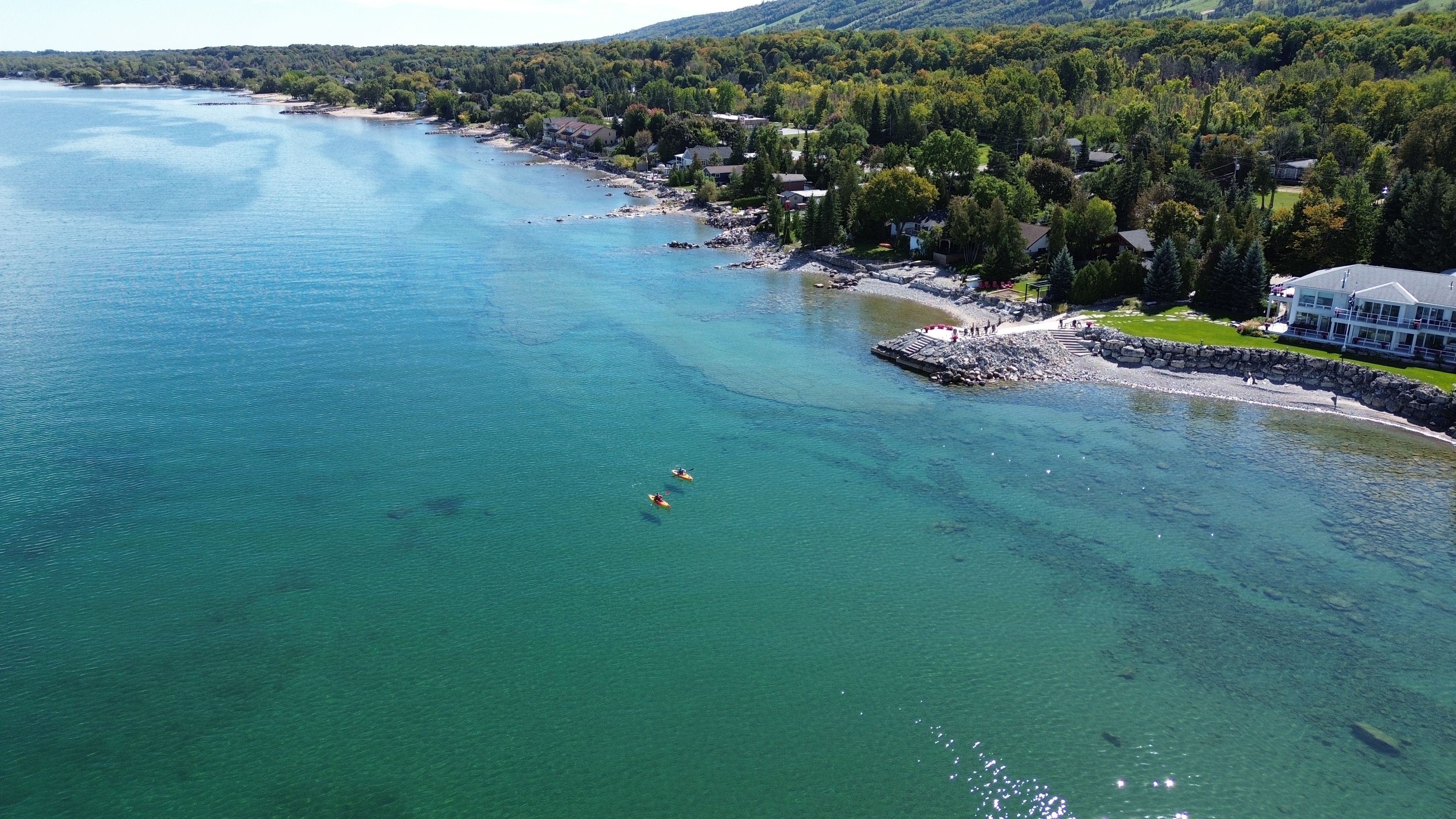 An aerial view of a coastal area with clear water, a rocky shoreline, and a mix of residential buildings and greenery.