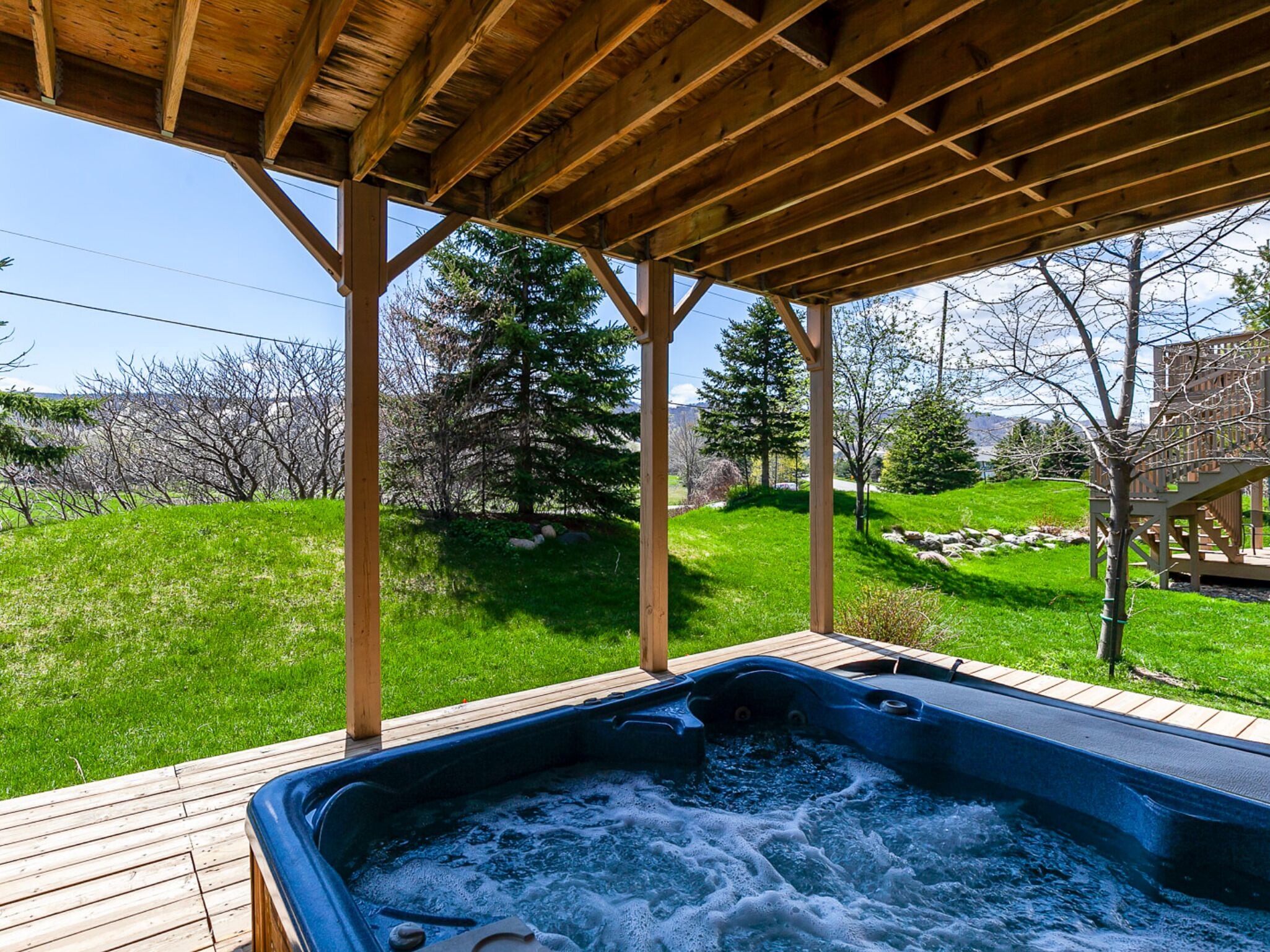 A wooden deck with a hot tub under a pergola, surrounded by a grassy area and trees.