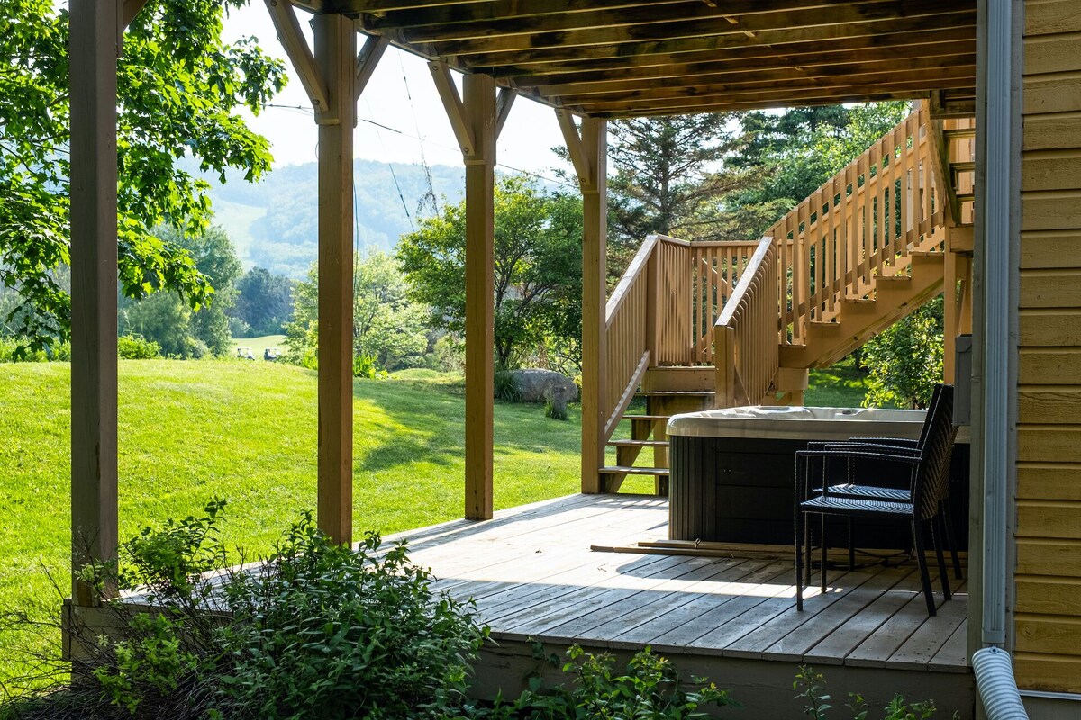 Une terrasse en bois avec un jacuzzi et des chaises, offrant une vue sur un paysage verdoyant et des collines.