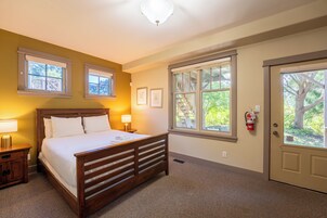 Habitación de hotel con cama de madera, dos lámparas de noche, una ventana con vistas a árboles y una puerta con extintor.