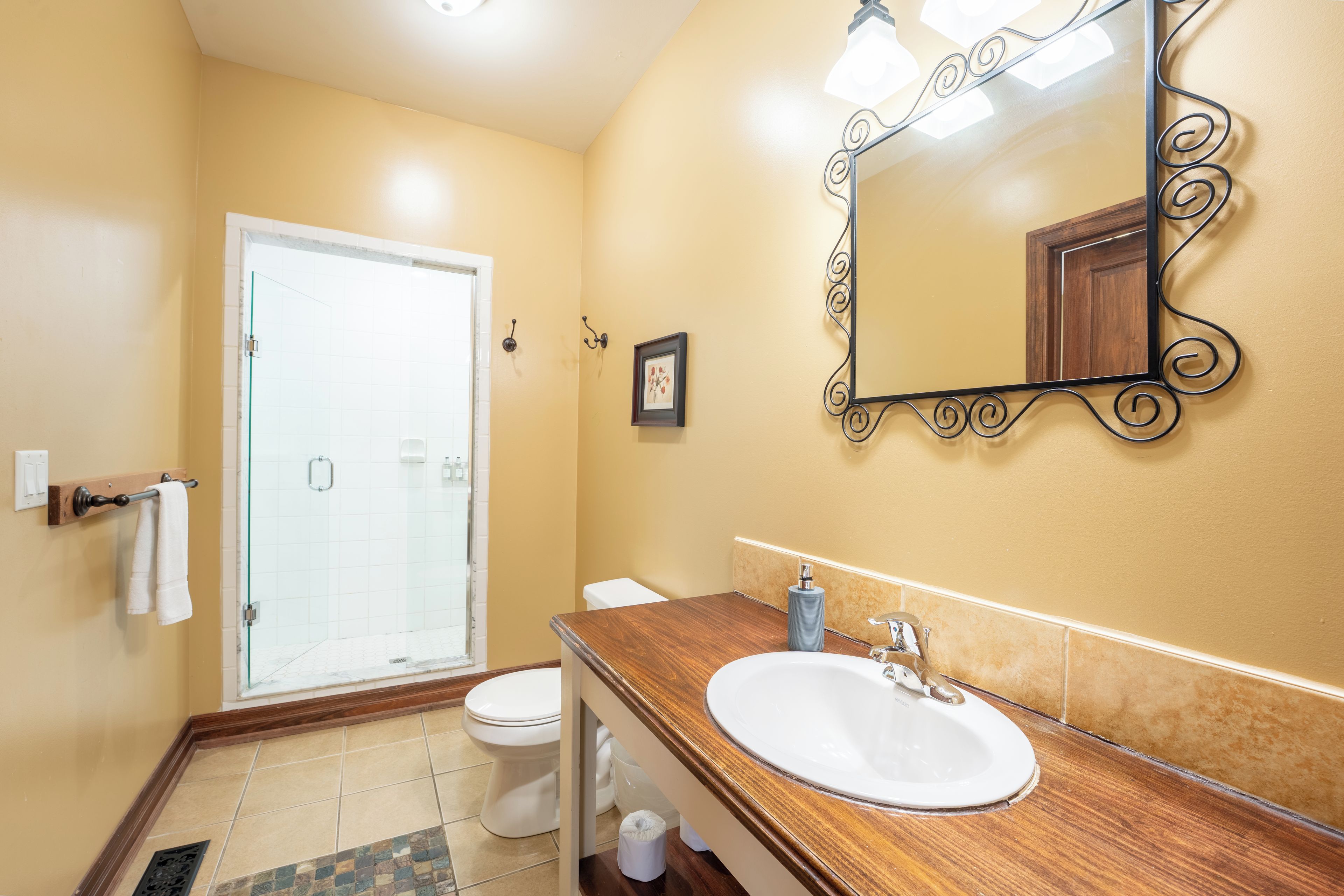 A bathroom with a wooden vanity, a white sink, a mirror with a decorative frame, a toilet, and a glass shower enclosure.