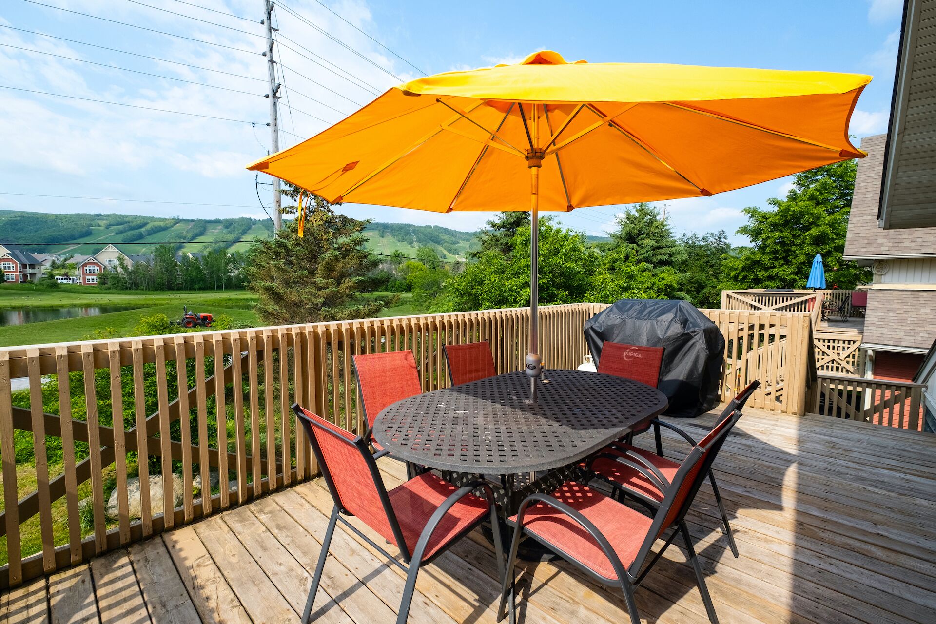 Een houten terras met een ronde tafel en stoelen, een gele parasol en een barbecue.