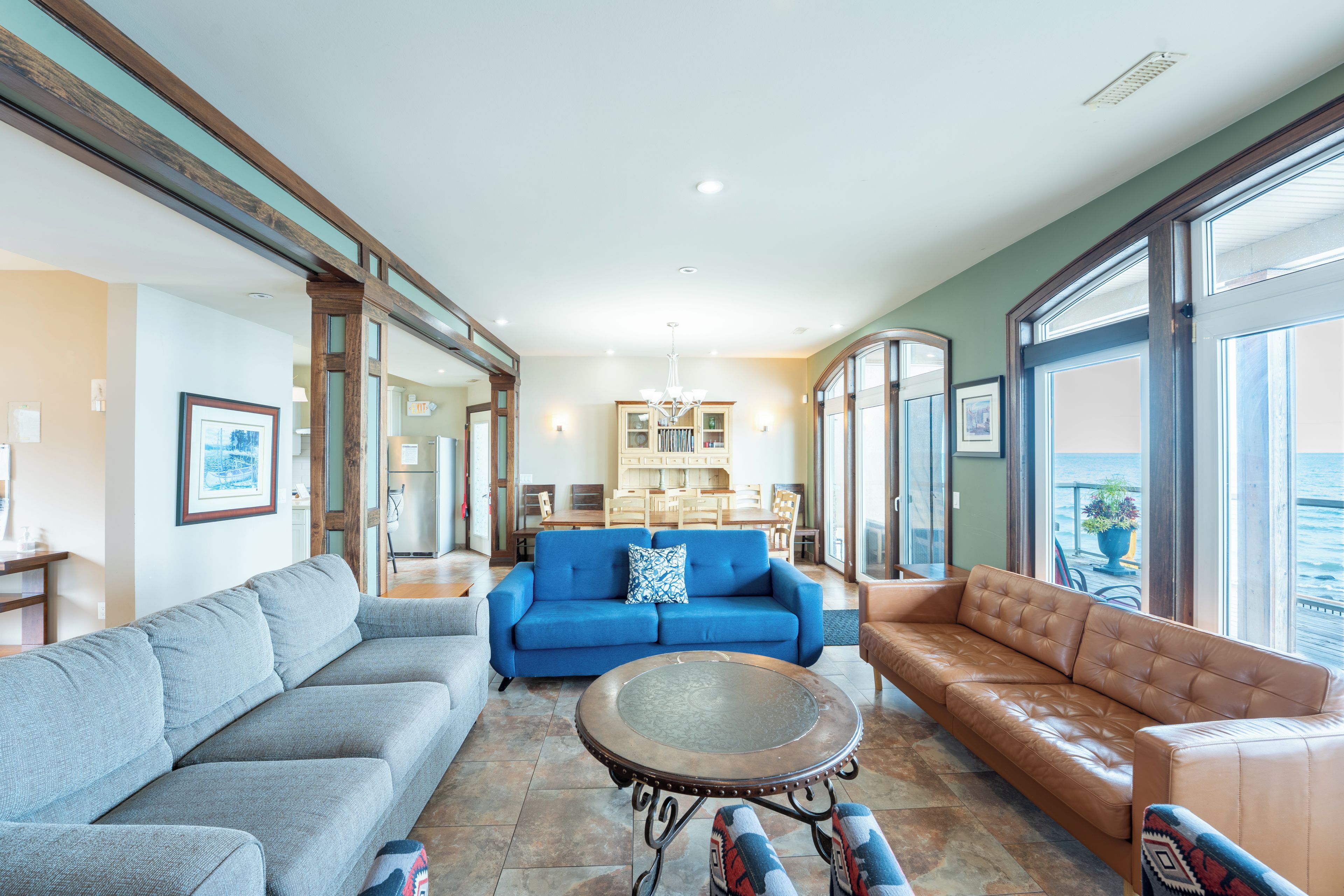 A spacious living area with a blue sofa, a brown leather sofa, and a grey sofa. There is a dining area in the background with a chandelier.