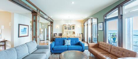 A spacious living area with a blue sofa, a brown leather sofa, and a grey sofa. There is a dining area in the background with a chandelier.