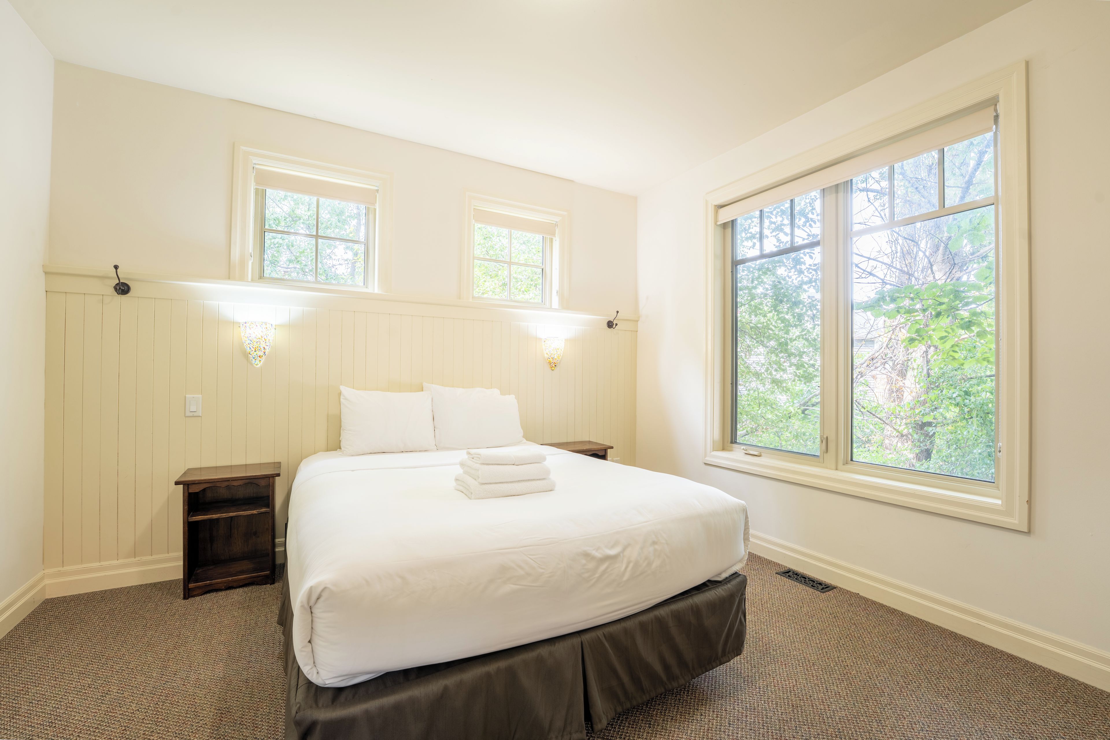 Un dormitorio con una cama grande, dos mesitas de noche, una lámpara de noche, una ventana con vistas a árboles y luces fijadas en la pared.