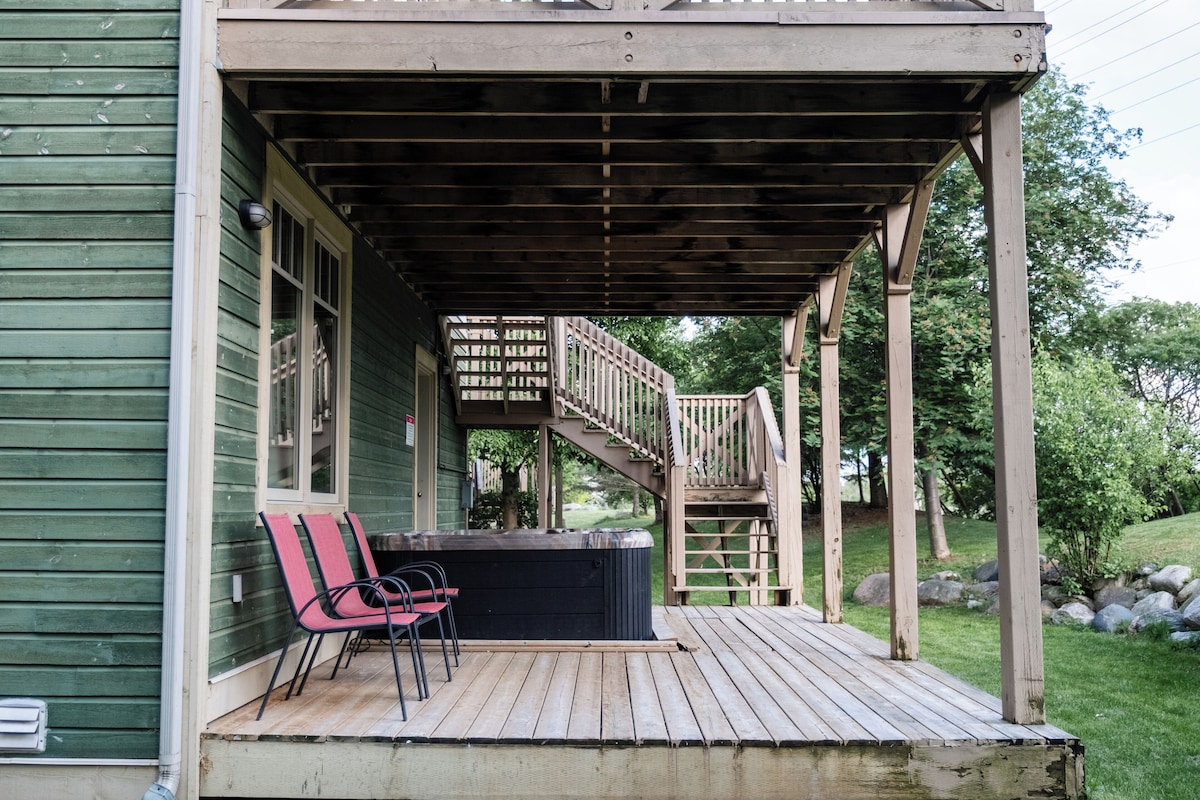 Une terrasse en bois avec un jacuzzi et des chaises, une véranda et un escalier menant à une autre terrasse.