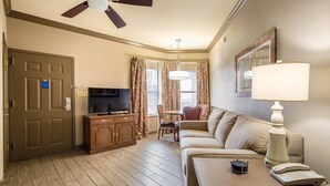 A hotel room with a television, a sofa, a dining table with chairs, a ceiling fan, and a lamp.