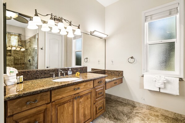 A bathroom with a granite countertop, wooden cabinets, a large mirror, and a window with frosted glass.