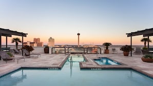 A rooftop pool with lounge chairs and potted plants, overlooking a city skyline at sunset.
