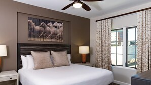 A hotel room with a large bed, two bedside tables with lamps, a ceiling fan, a window with curtains, and a wall-mounted painting.