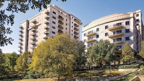 A multi-story residential building with balconies, a park with trees, and a water feature.