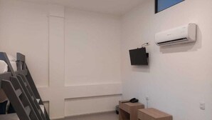 Une chambre avec un lit superposé, un climatiseur fixé au mur, une petite table de chevet en bois et une prise de courant.