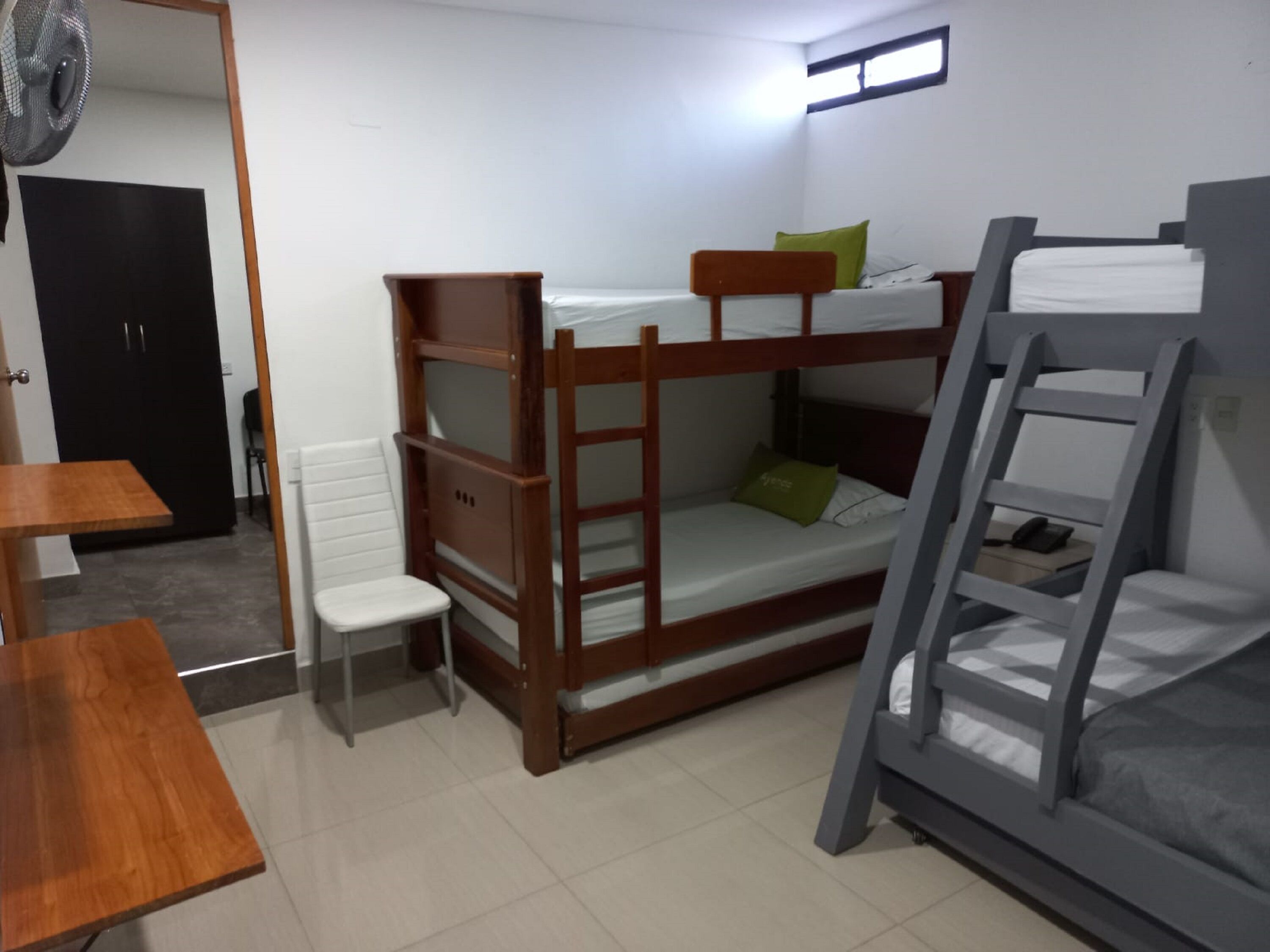 A dormitory room with bunk beds, a small desk, a chair, and a fan.