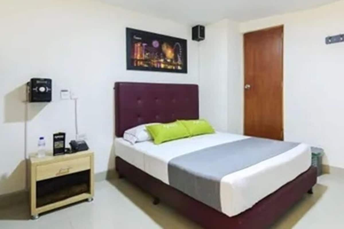 Een hotelkamer met een bed, nachtkastje, een speaker aan de muur en een deur.