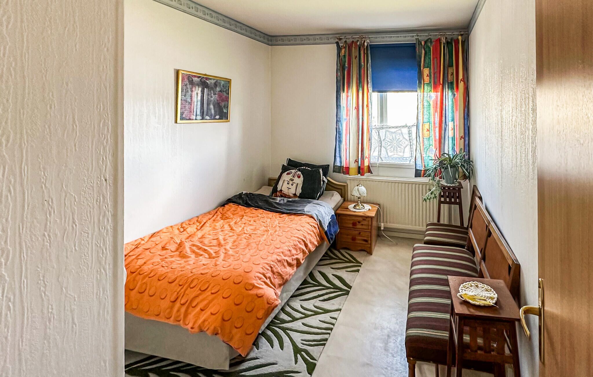 A bedroom with a bed, a bench, a nightstand, a window with curtains, and a radiator.