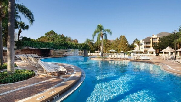 Relax by the resort pool surrounded by lounge chairs and umbrellas, with a wooden deck and palm trees in the background.