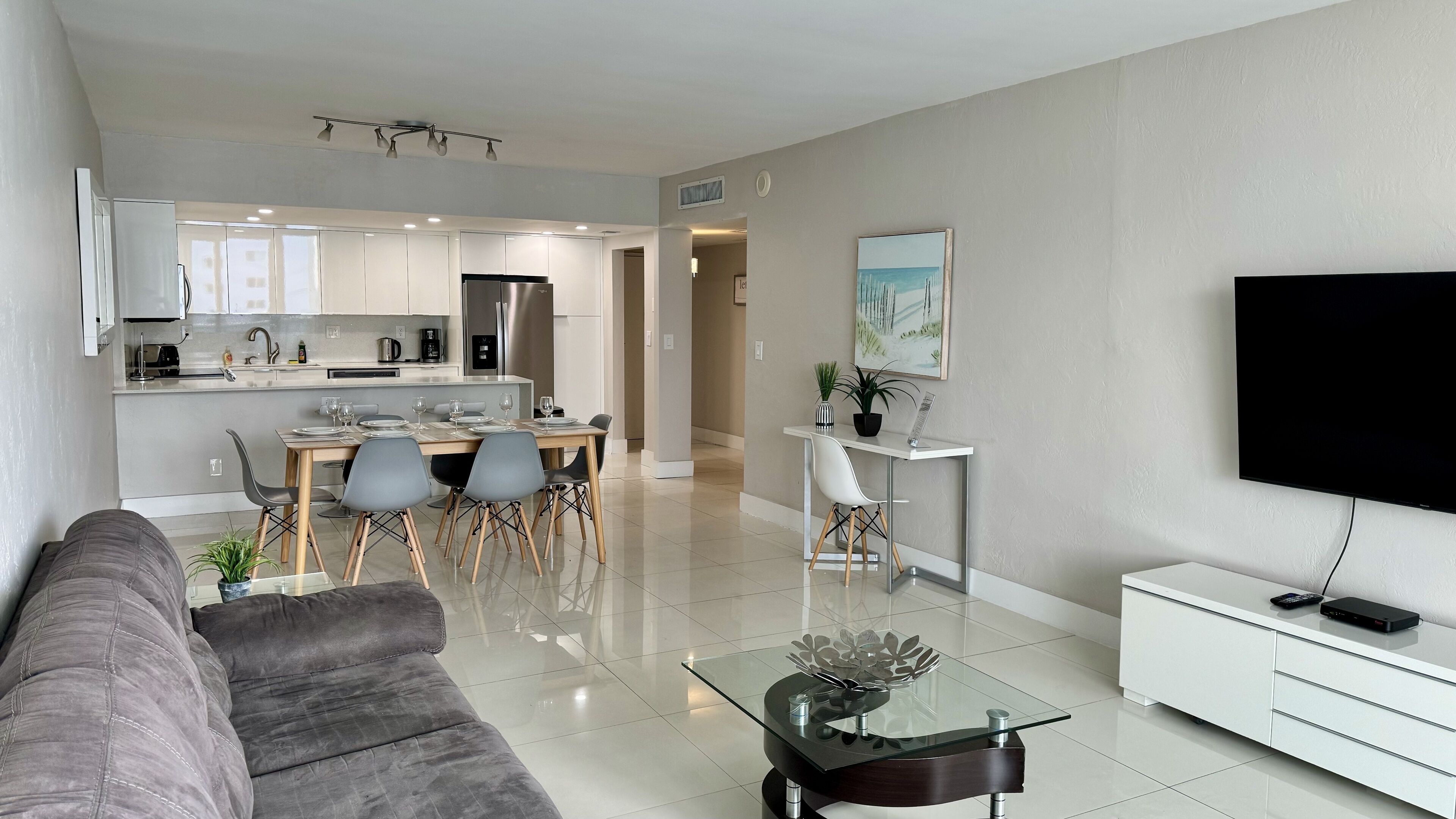 Un salon moderne avec un canapé gris, une table basse en verre et un téléviseur posé sur un meuble blanc.