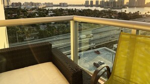 Un balcon doté de meubles en osier, d’une rambarde en verre et offrant une vue sur un paysage urbain avec des bâtiments et une étendue d’eau.
