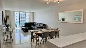 Un salon moderne avec un canapé en cuir noir, une table à manger pour quatre personnes et une grande fenêtre donnant sur des immeubles de grande hauteur.